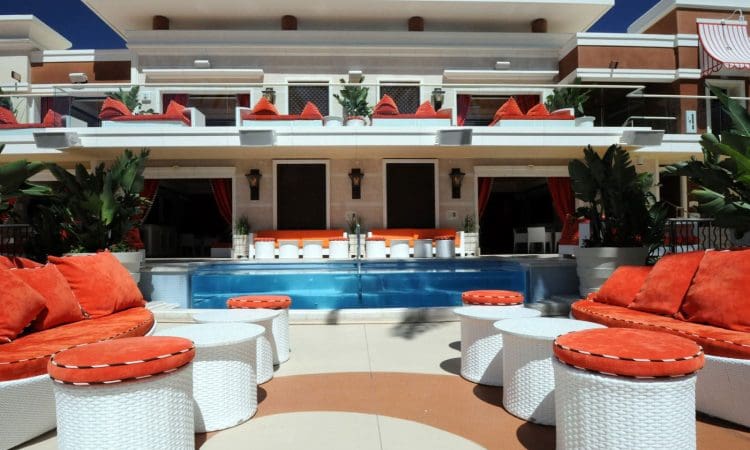 Outdoor lounge area with white wicker furniture and orange cushions, facing a small pool and a modern two-story building with balconies and cabanas.
