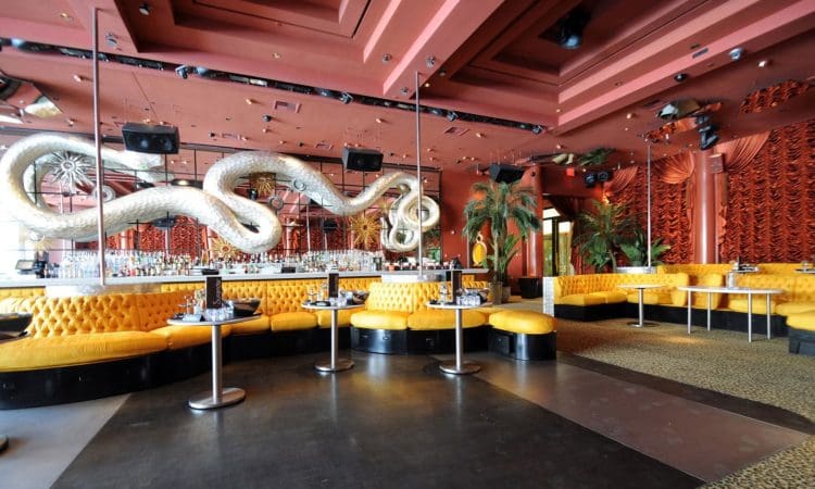 A stylish lounge with yellow tufted seating, small tables, mirrored walls, snake sculptures, and red patterned decor.