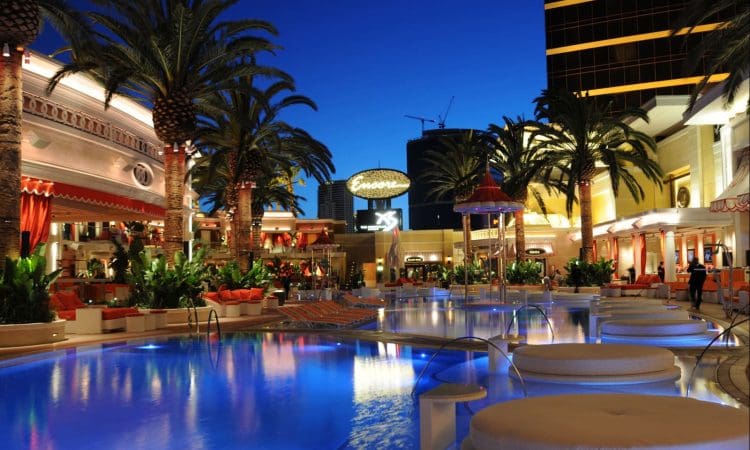 A brightly lit outdoor pool area at night with palm trees, lounge chairs, and the illuminated Encore hotel sign in the background.