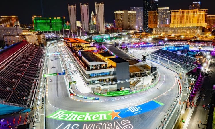 Aerial view of a nighttime Formula 1 racetrack in Las Vegas, surrounded by grandstands built by PENTA and lit-up city buildings, with "Heineken Las Vegas" painted on the track.