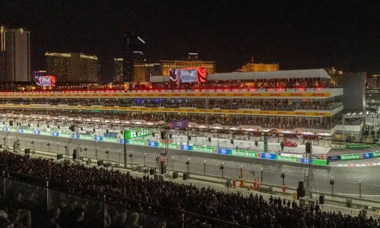 A nighttime view of a Formula 1 racetrack with brightly lit grandstands, packed audience, and city buildings illuminated in the background.