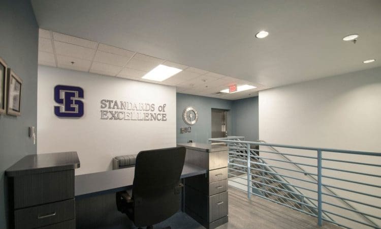 Reception desk area in an office hallway with "Standards of Excellence" sign on the wall, a clock, framed items, and metal railing beside stairs.