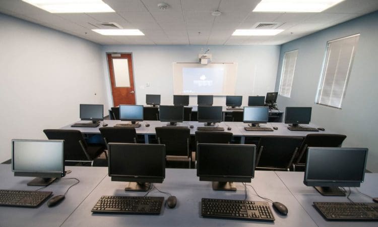 A computer lab with several rows of desks, each equipped with monitors, keyboards, and mice, facing a whiteboard and projector screen at the front of the room.