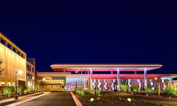 A modern, well-lit building with a wide entrance and landscaped surroundings is shown at night under a dark sky.