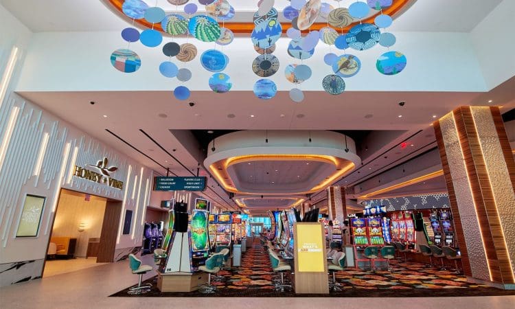Casino interior with colorful slot machines, modern lighting, suspended circular art pieces on the ceiling, and the entrance to a restaurant called Honey & Vine on the left.