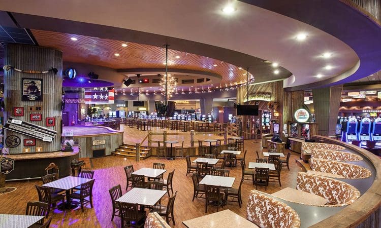 Spacious casino interior with empty dining tables, curved booths, slot machines in the background, wood flooring, and ambient lighting.