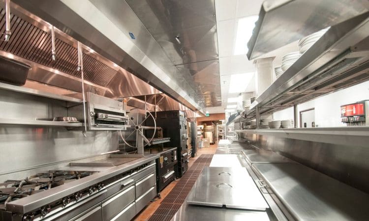 A commercial kitchen with stainless steel countertops, stoves, shelves, and various kitchen appliances, with clean, organized surfaces and dishes stacked on racks.