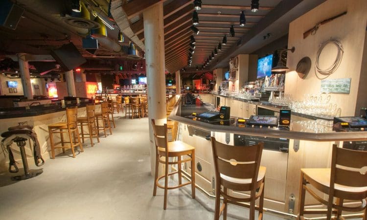 A spacious bar with wooden chairs and counters, glassware, wall-mounted décor, and illuminated shelves in a dimly lit setting.