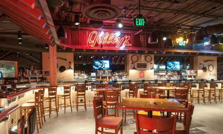 Interior of a spacious bar with wooden tables and chairs, a well-stocked bar, neon signs, and televisions mounted on the back wall.