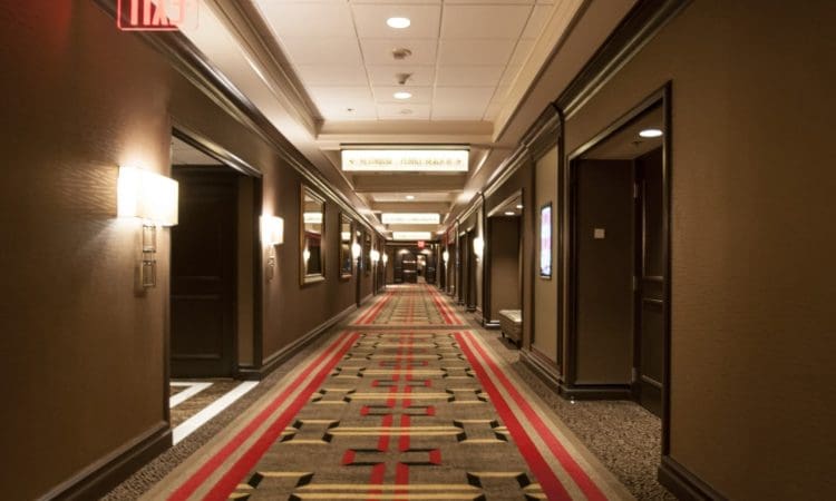 A hotel hallway with patterned carpet, wall lamps, room doors, and an illuminated exit sign on the ceiling.
