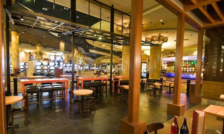 View from a restaurant area into a casino with slot machines, gaming tables, and neon lights, separated by glass panels and wooden beams.