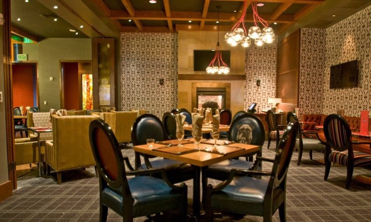 Modern restaurant dining area with dark wooden tables, black chairs, patterned wallpaper, contemporary lighting fixtures, and a fireplace in the background.