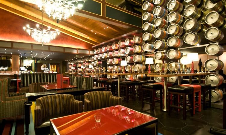 Modern restaurant interior with rows of metal beer kegs on the wall, red tables, striped chairs, bar stools, and decorative chandeliers.