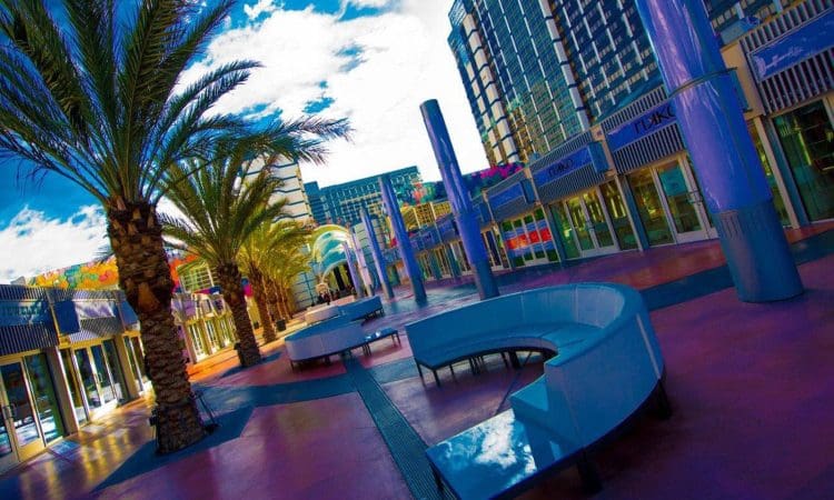 Outdoor plaza with curved benches, palm trees, and tall buildings under a partly cloudy sky. The image has vibrant, colorful lighting.