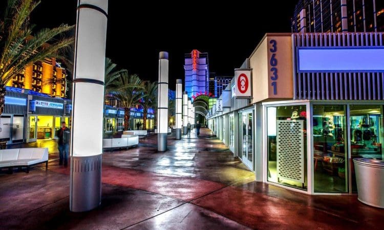 A brightly lit outdoor shopping walkway at night, lined with palm trees, illuminated pillars, and modern storefronts with glass windows.