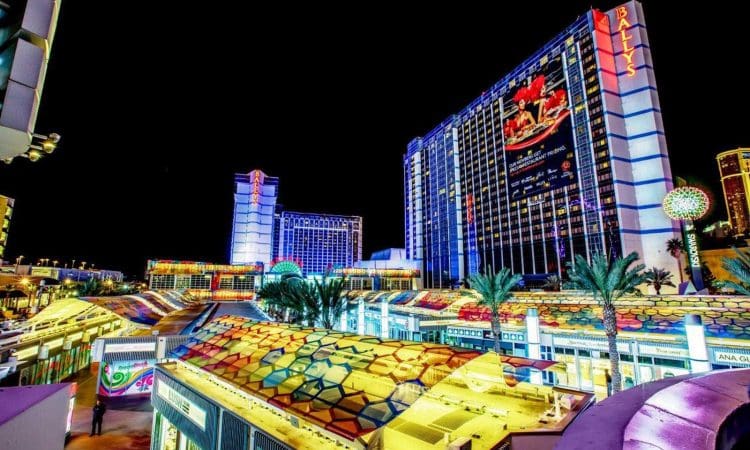 Brightly lit hotels and casinos, including Bally's, with neon lights and palm trees along a lively street at night in Las Vegas.