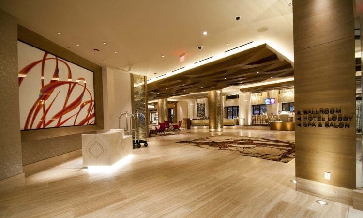 Modern hotel lobby with beige flooring, abstract wall art, luggage cart, and signs directing to ballroom, hotel lobby, spa, and salon. Reception desk and seating area visible.