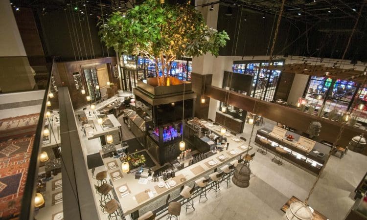 A modern restaurant interior with a central bar, high ceilings, a large tree feature, and surrounding seating, viewed from above.