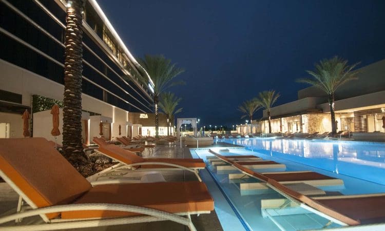 Outdoor swimming pool at night with orange lounge chairs, palm trees, and modern buildings with lights reflecting on the water.