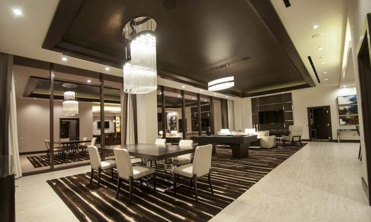 Modern dining and lounge area with large windows, dark ceiling, contemporary chandeliers, and cream-colored chairs around rectangular tables on striped rugs.