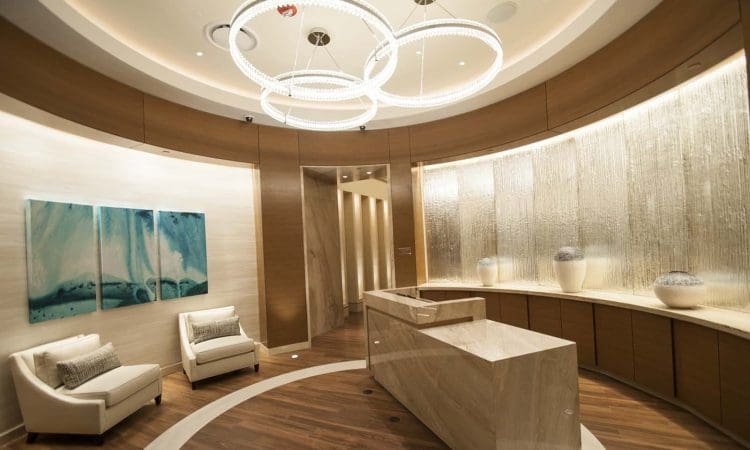 Modern lobby interior with marble reception desk, circular light fixtures, two white armchairs, abstract artwork, and a wall with water feature and decorative vases.