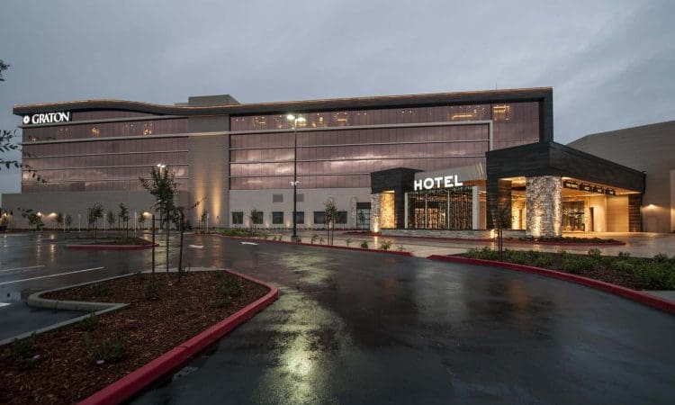 Modern hotel building with large glass windows, labeled "HOTEL" in illuminated letters, and an adjacent structure with the name "GRITON" at dusk. Wet pavement in front.