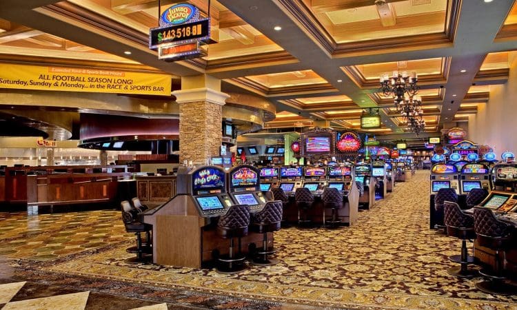 Rows of illuminated slot machines line a carpeted casino floor under ornate ceiling lights, with a sports bar visible in the background.