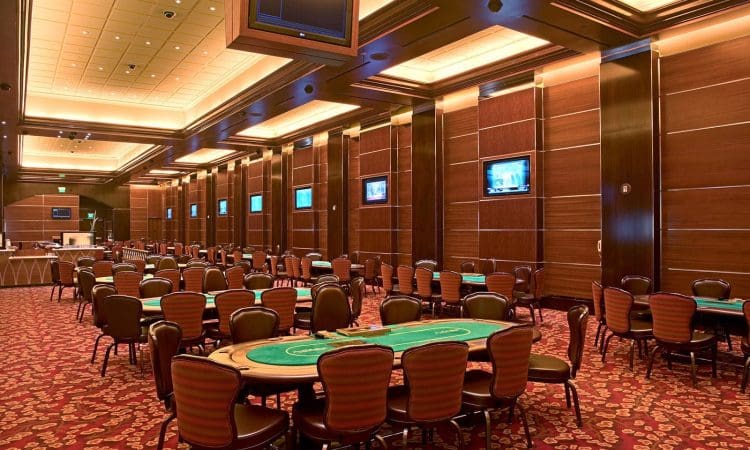 Empty casino poker room with green card tables, rows of chairs, flat-screen TVs on wood-paneled walls, and patterned red carpeting under warm ceiling lights.