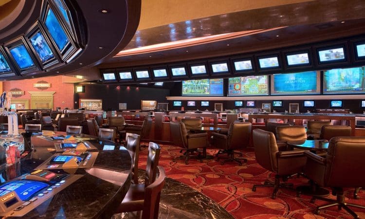 A casino sportsbook area with rows of chairs, electronic betting terminals, and multiple large screens displaying sports events and odds.