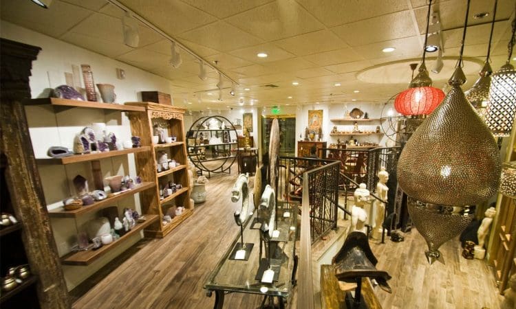 A well-lit store interior displays shelves of crystals, statues, and decorative items on wood flooring, with large ornate lamps hanging from the ceiling.