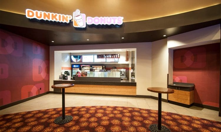 Dunkin' Donuts storefront with counter, menu boards, and coffee machines; two standing tables and patterned carpet in the foreground.