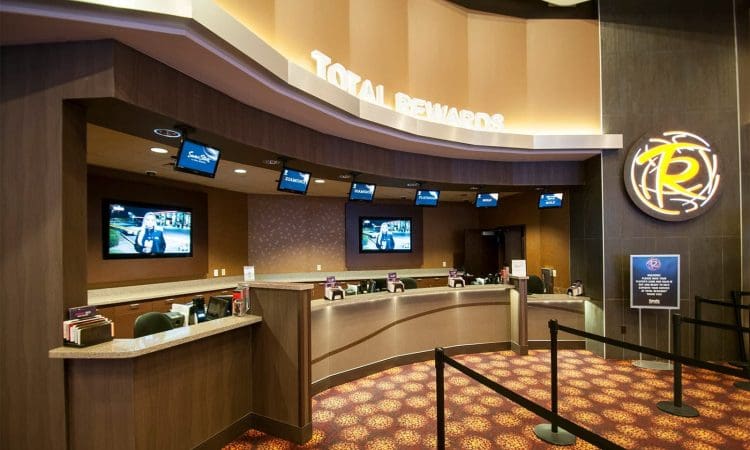 A casino rewards desk with multiple service windows, computer screens, overhead monitors, and a rope barrier on a patterned carpet.