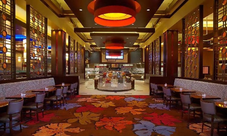 A modern restaurant interior with patterned carpet, booth and table seating, decorative panels, and an open buffet area in the background under warm ceiling lights.
