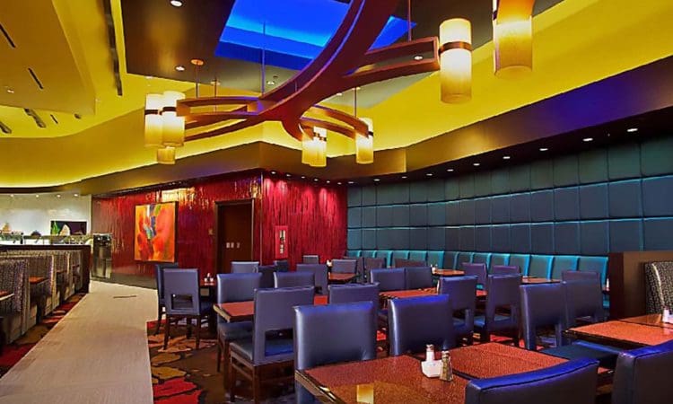 Modern restaurant interior with blue padded booths, dark wooden tables, and contemporary lighting fixtures under a yellow ceiling with a blue skylight. No people are present.