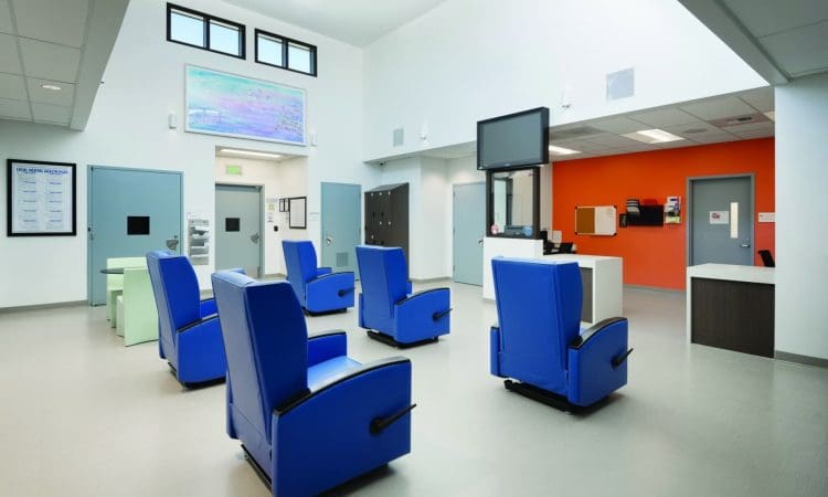 A brightly lit waiting area with several blue reclining chairs, a wall-mounted TV, and a reception desk against an orange accent wall.