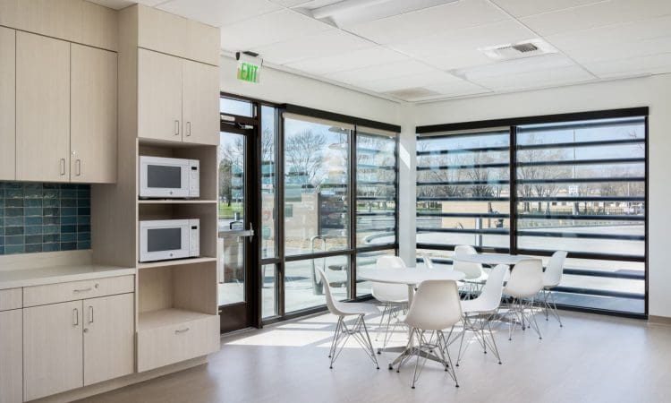 Modern break room with white chairs and round tables near large windows, built-in cabinetry, two microwaves, and a view of trees outside.