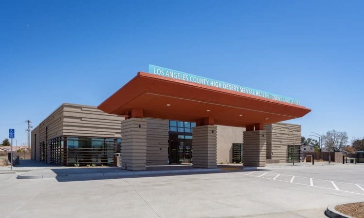 Single-story building with a prominent orange entrance canopy labeled "Los Angeles County High Desert Mental Health Urgent Care" and an empty parking lot in front.
