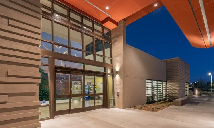 Modern building entrance at night with large glass doors, textured walls, and bright overhead lights illuminating the walkway.