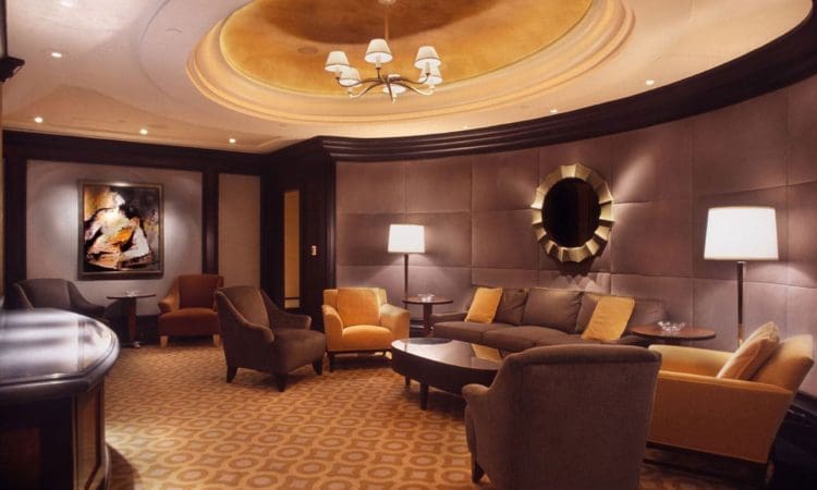 Modern lounge with armchairs, sofas, lamps, a circular mirror, abstract wall art, and a round ceiling detail with a chandelier and yellow lighting.