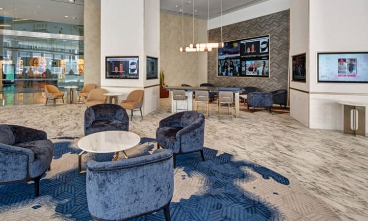 Modern hotel lobby with blue and beige seating, round tables, wall-mounted screens, and marble-patterned floors. Large windows provide natural light.