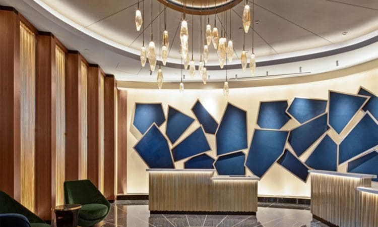 Modern hotel lobby with geometric blue artwork on the wall, gold accent lighting, hanging glass pendant lights, and green velvet chairs on black marble flooring.