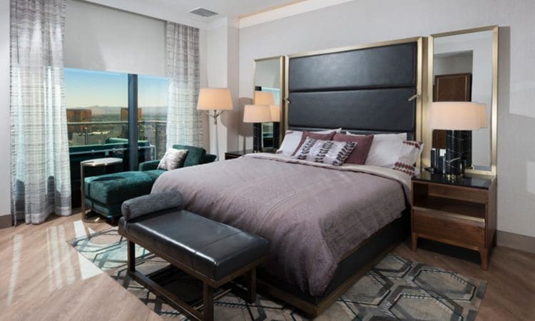 Modern hotel room with a large bed, two bedside tables with lamps, a green lounge chair by the window, and a patterned area rug on wood flooring.
