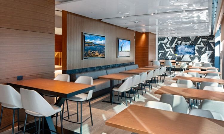 Modern cafeteria with wooden tables, white chairs, wall-mounted landscape photos, wood paneling, and a geometric black-and-white accent wall at the back.