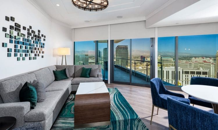Modern living room with gray sectional sofa, teal accents, abstract wall art, large windows, city skyline view, and a round dining table with blue chairs.