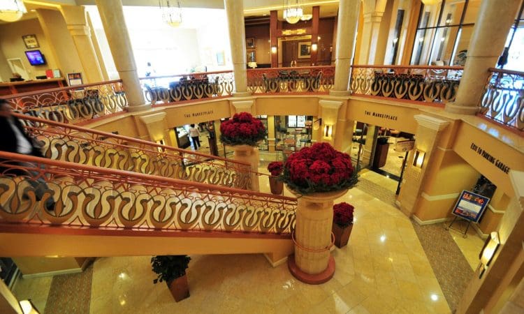 Interior view of a hotel lobby with grand staircases, decorative railings, marble floors, large columns, and potted red flowers arranged throughout the space.