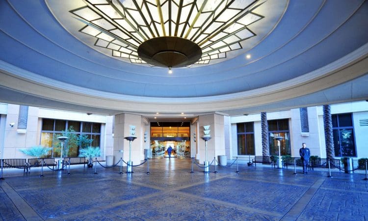 Covered entrance to a building with a large decorative ceiling light, textured flooring, and glass doors leading inside. Benches and plants line the entryway.