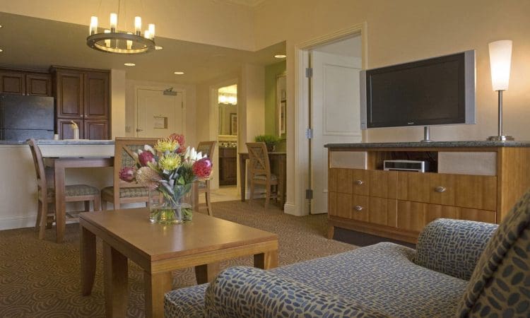 A hotel suite with a patterned armchair, wooden coffee table with a flower arrangement, TV on a dresser, dining area, and kitchenette in a warmly lit, neutral-toned space.