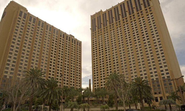 Two tall, beige high-rise buildings with vertical dark window columns stand side by side, surrounded by palm trees and a street with a passing car in front.