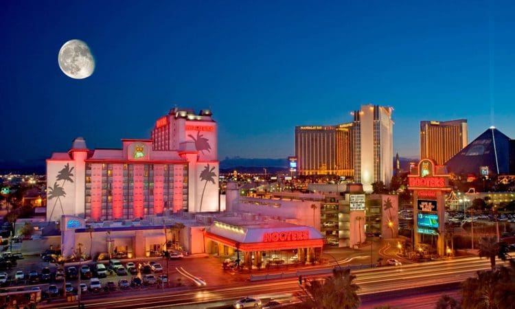 Night view of Las Vegas Strip with illuminated buildings, including a Hooters hotel and casino, traffic light trails, and a large moon visible in the sky.