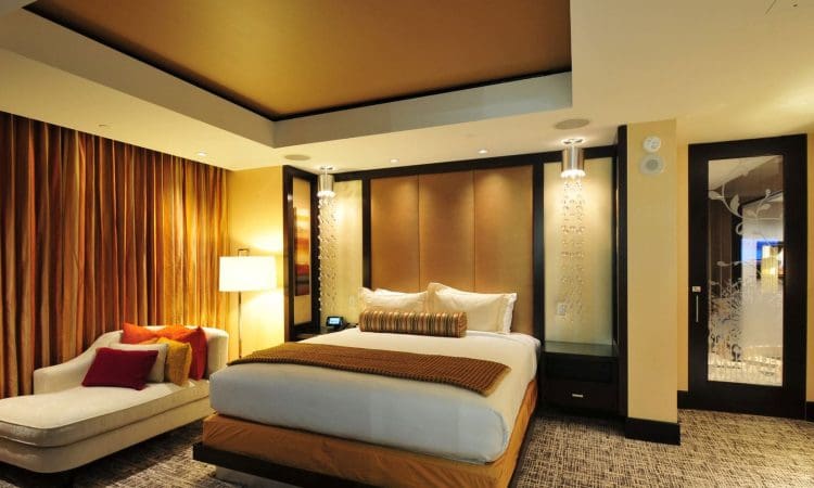 Modern hotel room with a king bed, padded headboard, chaise lounge, warm lighting, brown and gold accents, and floor-to-ceiling curtains.